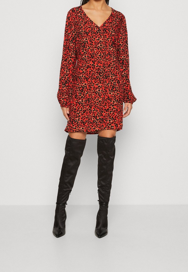 Femme portant une robe longue à manches longues imprimée léopard rouge et noire, assortie à des bottes noires à talons au-dessus du genou, debout devant un fond uni.