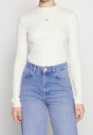 Personne portant un haut blanc à manches longues côtelé avec un patch logo Calvin Klein et un jean bleu clair taille haute.