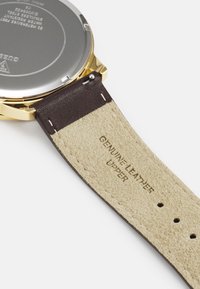Goldfarbene Uhr mit silbernem Edelstahlrücken, ausgestattet mit einem dunkelbraunen Echtlederarmband mit beiger Unterseite und geprägtem Text.