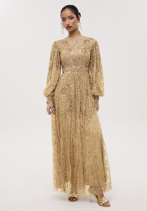 Femme portant une longue robe dorée avec broderies en sequins, manches longues bouffantes et sandales à talons hauts dorées avec détails floraux.