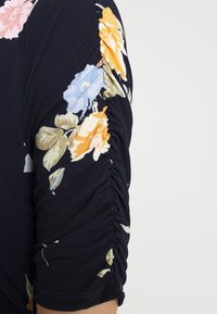 Navy blue vaatehiha, jossa on oransseja, sinisiä ja beigejä kukkakuoseja. Hihaan kulkee pystysuora rypytetty sauma.