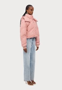 En pufferjacka i pastellrosa, med hög krage, dragkedja och ribbade manschetter. Kombinerad med ljusblå vida jeans och sandaler med remmar.