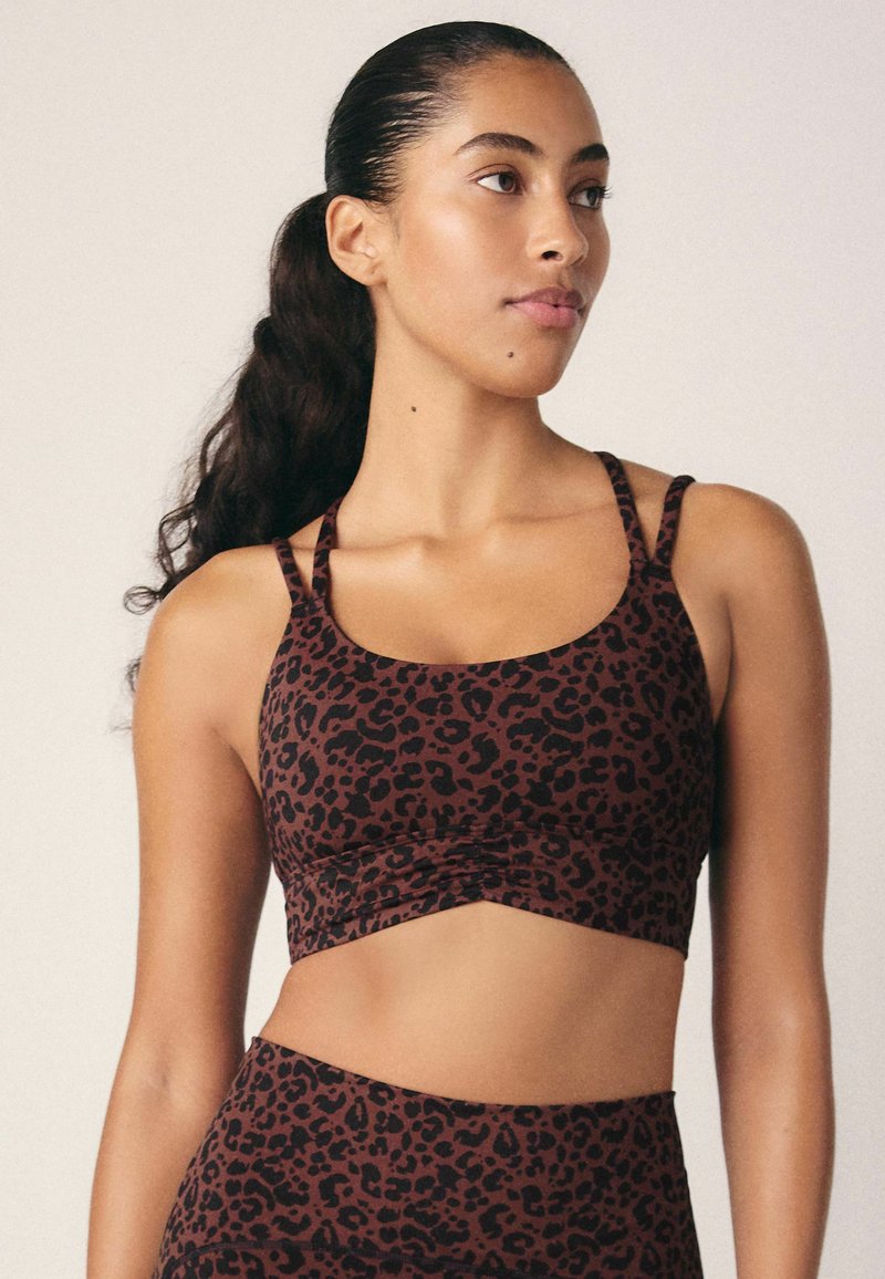Sujetador deportivo marrón con un diseño de leopardo negro, con tirantes finos y un corte cropped. El material parece suave con un ligero brillo.