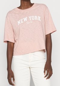 Lyserød cropped t-shirt med korte ærmer, der har et hvidt "NEW YORK" print og blødt, let tekstureret stof.