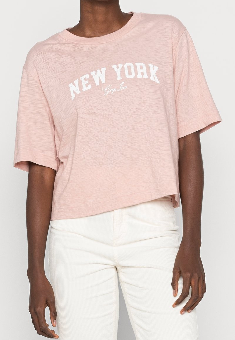 Lyserød cropped t-shirt med korte ærmer, der har et hvidt "NEW YORK" print og blødt, let tekstureret stof.