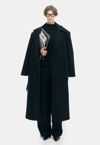 Zwarte oversized jas met een brede kraag, gedragen over een zwarte trui en broek. Houdt documenten vast, benadrukt minimalistisch ontwerp en monokrome textuur.