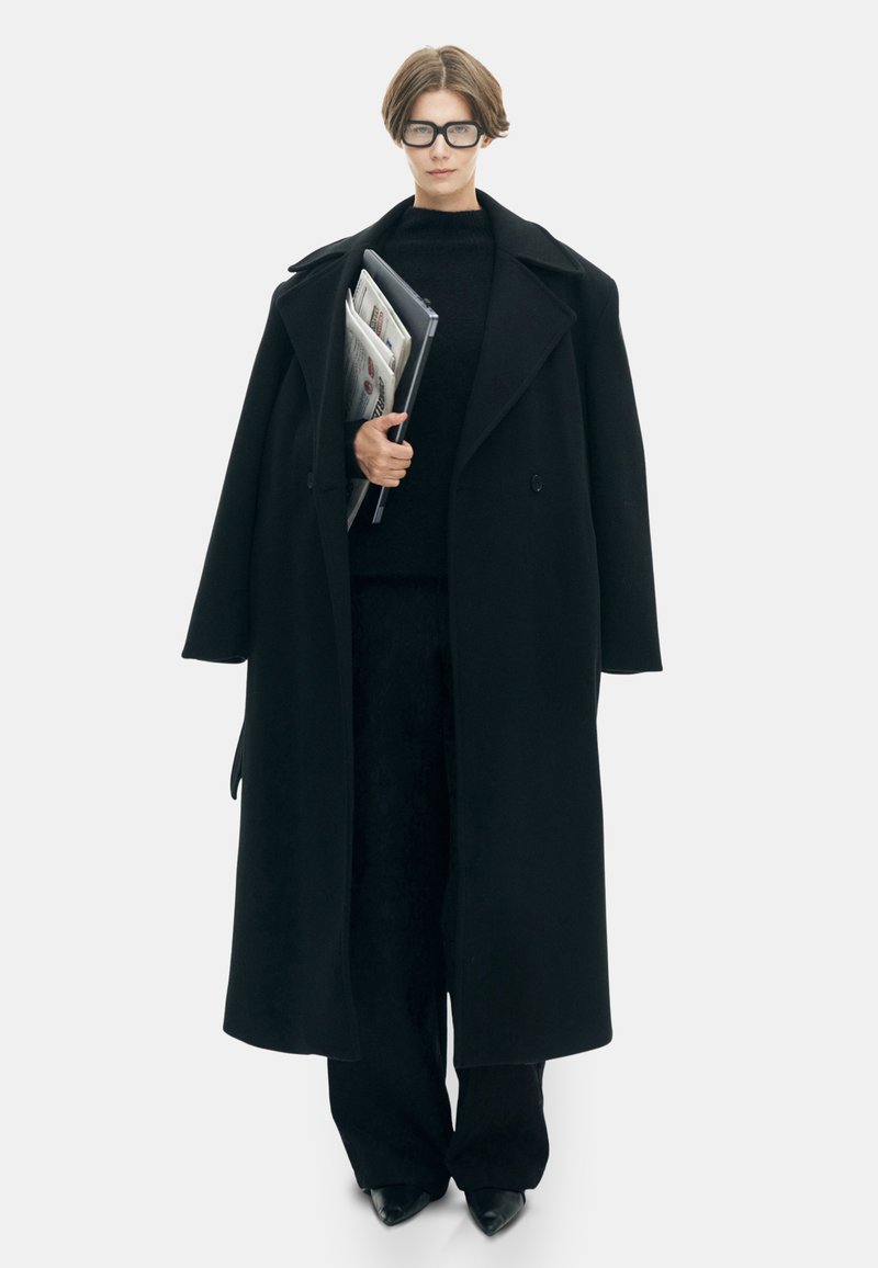 Zwarte oversized jas met een brede kraag, gedragen over een zwarte trui en broek. Houdt documenten vast, benadrukt minimalistisch ontwerp en monokrome textuur.