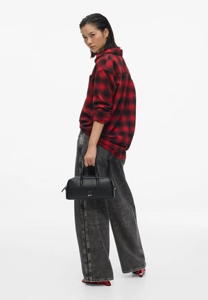 Femme portant une chemise à carreaux rouge et noire, jean noir délavé à jambes larges, talons rouges, tenant un petit sac à main noir sur un fond blanc.