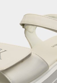 Gros plan sur une sandale en cuir beige avec une bride ajustable et des coutures visibles sur la partie supérieure.