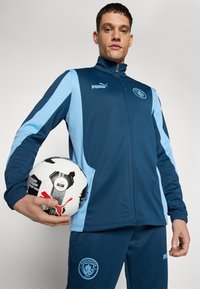 Mornarsko in svetlo modro trenirko z zadrgo na jakni in hlačah, ki vsebuje logotip Puma in emblem Manchester City. Drži črno-belo nogometno žogo.