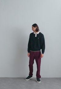 Hanorac negru cu guler și logo alb, asociat cu pantaloni burgundy. Încălțăminte neagră cu talpă albă. Design minimalist.