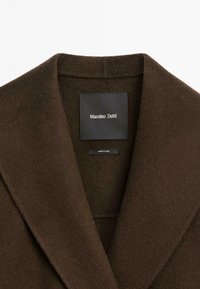 Hnědý vlněný kabát s klopovým límcem. Měkký, strukturovaný materiál. Etiketa nese nápis "Massimo Dutti" s menší nálepkou uvádějící "Vyrobeno v Číně."