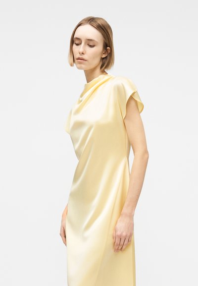 Γυναίκα με κοντά ξανθά μαλλιά που φοράει φορεματίνα από απαλό κίτρινο σατέν, στέκεται μπροστά σε απλό ανοιχτό γκρι φόντο, κοιτάζοντας προς τα κάτω.