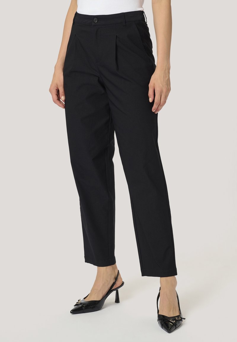 Femme portant un pantalon noir taille haute à jambes droites avec des plis, assorti à des talons slingback noirs à bouts pointus et un haut blanc.