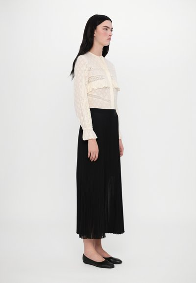 Blusa marfil con encaje en los bordes y acentos con volantes, combinada con una falda plisada negra y zapatos planos negros. El fondo neutro resalta el conjunto.