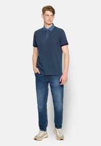 Tengerészkék polo ing chambray gallérral, rövid ujjú és hímzett logóval. Kék farmernadrággal és több színárnyalatú sportcipőkkel viselve.