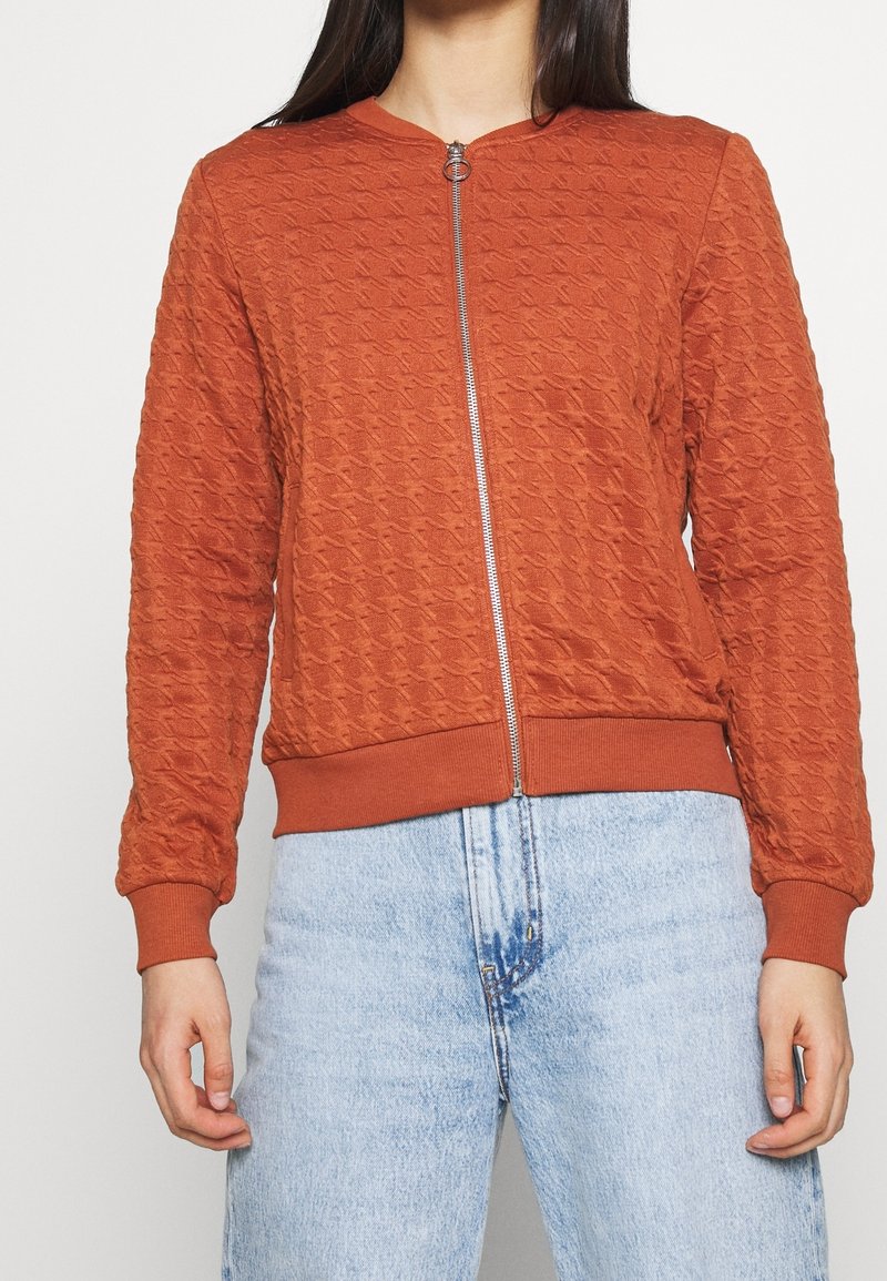 Femme portant une veste zippée texturée couleur rouille orangée avec poignets et ourlet côtelés, associée à un jean taille haute bleu clair.