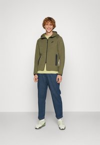 Olive green zip-up hoodie with black accents, kangaroo pockets, paired with a light yellow shirt, blue denim pants, and white sneakers.