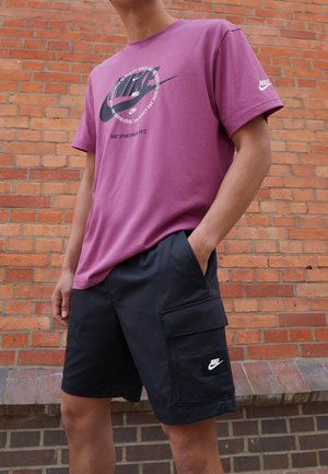 Fioletowy t-shirt Nike z nadrukiem logo, połączony z czarnymi szortami cargo z przednią kieszenią i naszywką z logo. Gładki kolor i luźny krój.