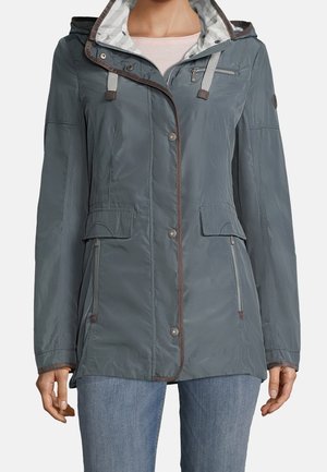 Vrouw die een grijze capuchonjas draagt met knoop- en ritssluitingen, klepzakken en een blauwe spijkerbroek, staande tegen een egale achtergrond.