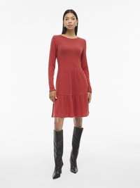 Červené žebrované šaty s dlouhým rukávem a rozšířenou sukní, se svislou strukturou. Doplněné vysokými černými lodičkami.