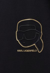 Μαύρο t-shirt με γραφικό ενός προσώπου με γυαλιά ηλίου σε χρυσό και μαύρο περίγραμμα, με το κείμενο "KARL LAGERFELD" κάτω από το σχέδιο.