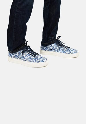 Canvas sneakers met een blauw-wit bloemenpatroon, met witte zolen en donkere veters, gecombineerd met donkere denim jeans.