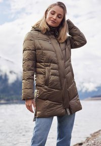 Olivenfarbene gepolsterte Jacke mit Kapuze, diagonal gestepptem Design, seitlichen Taschen und einem leichten Glanz. Über Jeans getragen.