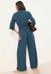 Blauer Einteiler mit langen Ärmeln, tailliert mit Gürtel, weit geschnittenen Beinen und Kragen, kombiniert mit einer braunen Wildleder-Handtasche. Glatte Textur.