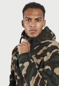 Chaqueta de forro polar con diseño de camuflaje en tonos terrosos de verde, marrón y crema. Presenta un cuello alto y cierre de cremallera con cordones. Textura suave.