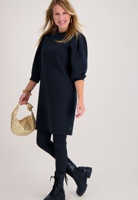 Marineblauwe oversized jurk met ballonmouwen, gecombineerd met zwarte legging en zwarte veterlaarzen. Houdt een gouden metalen tas vast.