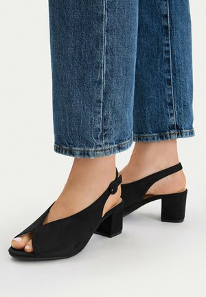 Pies con tacones de bloque slingback de ante negro combinados con jeans de mezclilla azul recortados sobre un fondo blanco.