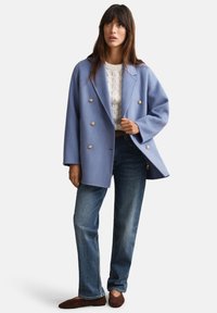 Cappotto doppiopetto in lana azzurro chiaro con bottoni dorati, indossato sopra un maglione bianco lavorato a maglia; abbinato a jeans a gamba dritta e ballerine marroni.