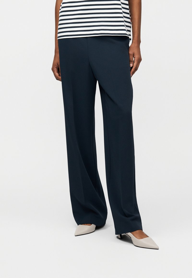 Γυναίκα με μαύρο παντελόνι φαρδιάς γραμμής, λευκά παπούτσια με χαμηλό τακούνι και μυτερή μύτη, και μαύρο-άσπρο ριγέ κοντομάνικο τοπ, στέκεται μπροστά σε λευκό φόντο.