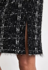 Robe texturée noir et blanc avec un ourlet à franges et une fente sur le côté, présentant un motif tacheté et une apparence douce et tissée.