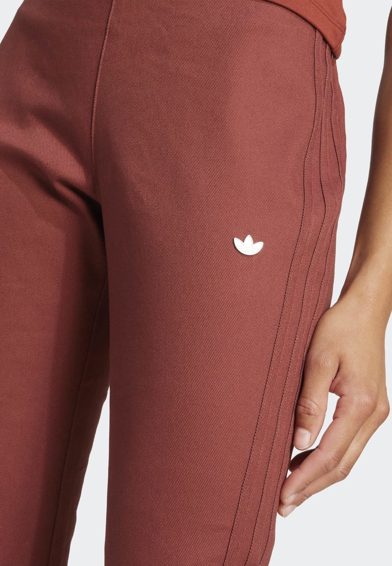 Gros plan sur une personne portant un pantalon Adidas couleur rouille avec le logo trèfle sur la cuisse et des bandes latérales cousues visibles.