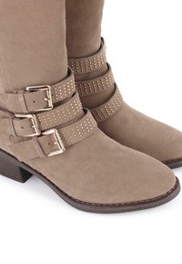 Botas de tobillo de ante en tono taupe, con tres correas decorativas que presentan tachuelas en tono dorado y hebillas de metal, además de un bajo tacón de madera.