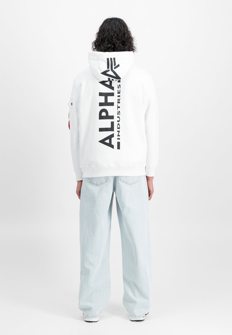 Personne aux cheveux bouclés portant un sweat à capuche blanc avec le texte noir « Alpha Industries » dans le dos et un jean large bleu clair, vue de dos.