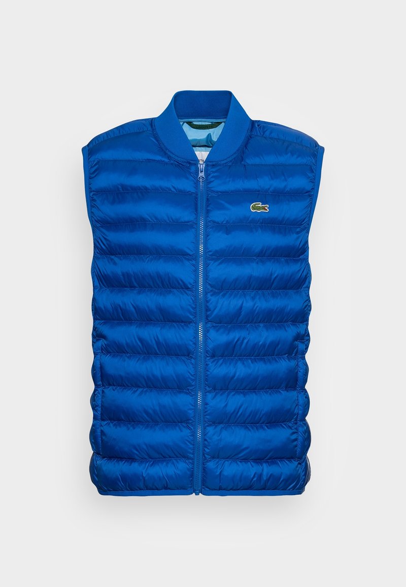 Veste matelassée bleue sans manches avec fermeture éclair à l'avant et petit logo vert brodé sur le côté gauche de la poitrine.