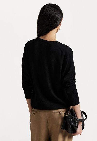 Black knit sweater with a round neckline and raglan sleeves, paired with tan pants and a small black handbag held in the right hand.