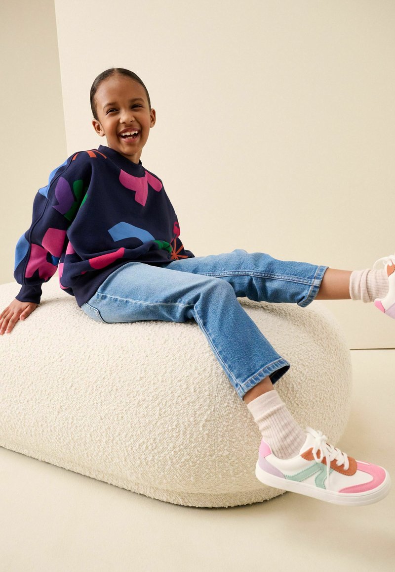 Niño sonriente sentado sobre un cojín blanco con textura, vestido con una sudadera colorida, vaqueros azules, calcetines blancos y zapatillas rosas y blancas.
