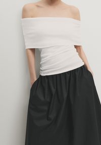 Valkoinen off-the-shoulder-toppi, jossa on teksturoitu kangas, istuva vartalo ja kerrosrakenne, yhdistetty mustan, runsaan hameen kanssa, jossa on taskut.