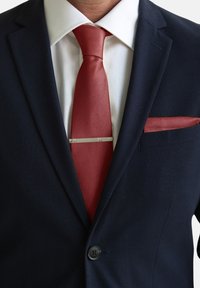 Chaqueta de traje en azul marino con una corbata roja que presenta un patrón texturizado y un pañuelo a juego. Clip de corbata plateado y detalle de botón negro visible.