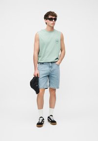 Giovane uomo con occhiali da sole, maglietta verde menta senza maniche, pantaloncini di jeans, sneakers nere, calzini bianchi, che tiene un cappello a righe nero in uno studio bianco.