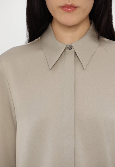 Light beige blouse made of smooth fabric, featuring a pointed collar, button closure, and a subtle sheen. Simple, clean design with no patterns.