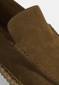Bruine suède instapper met een gestikt ontwerp, een platte zool en een gevlochten accent langs de rand. Gladde textuur en een eenvoudige silhouet.