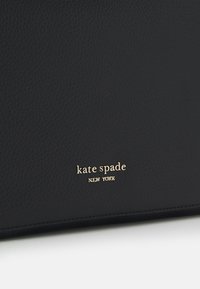 Superficie in pelle nera texturizzata con il logo dorato "kate spade NEW YORK" in rilievo vicino al bordo inferiore.