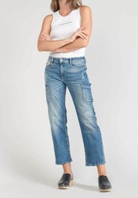 Mujer de pie con los brazos cruzados, vistiendo una blusa sin mangas blanca, pantalones cargo azules desgastados y zuecos negros, sobre un fondo neutral.