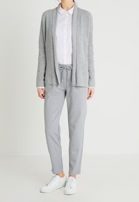 Hellgrauer Cardigan mit geripptem Design und offener Vorderseite, getragen über einem weißen Hemd mit Kragen. Kombiniert mit grauen Jogginghosen mit Kordelzug und weißen Sneakern.