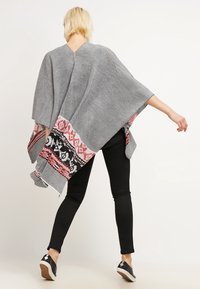 Grå strikket poncho med en farverig mønsterkant. Har V-hals, løs pasform og slids ved siderne, parret med sorte leggings og sko.